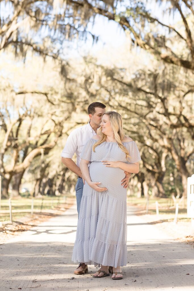 Soon-to-be parents stand in path covered by spanish moss trees during their Savannah maternity session as dad stands behind mom and holds her belly together with soon-to-be mom.