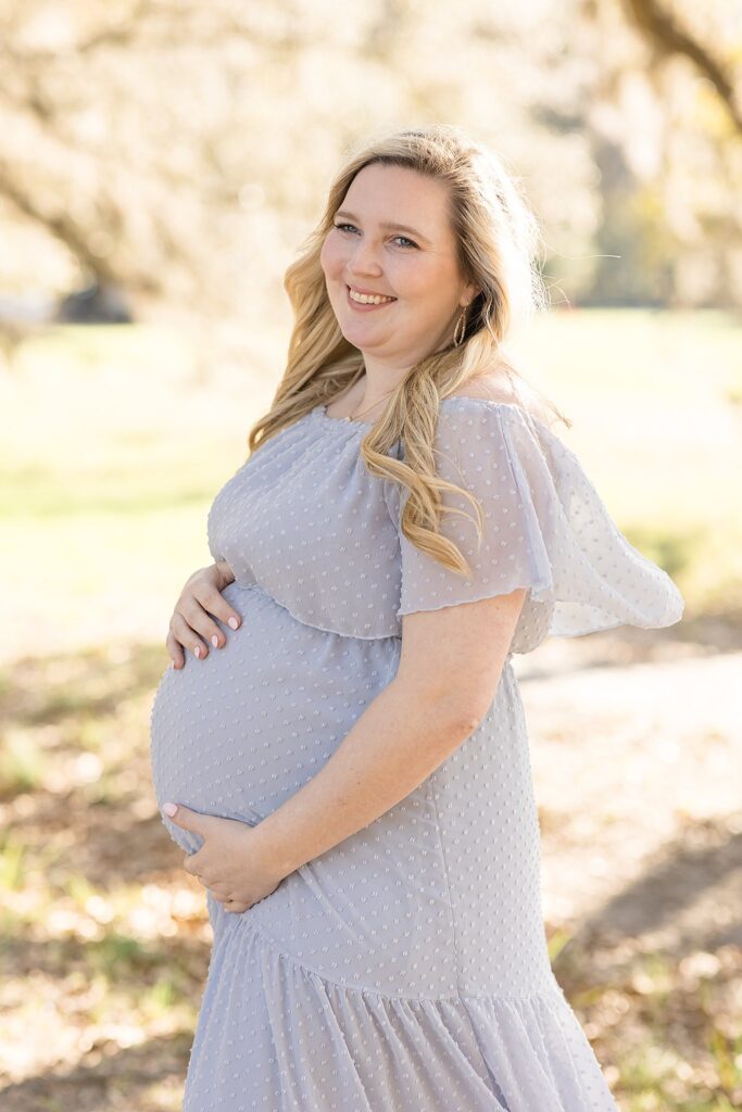 Gorgeous mom-to-be holds baby belly in her sheer, grey, eyelet dress as she smiles joyfully in the distance at her husband.