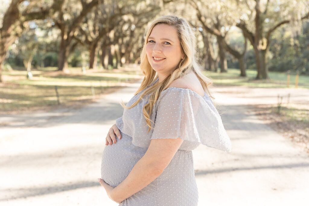 First time pregnancy with mom-to-be stands on a walkway at Wormsloe Historic Site during her Savannah maternity session with Wisp + Willow Photography Co.