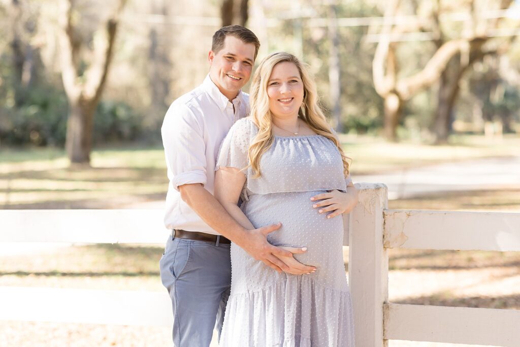 Husband holds wife's belly as he leans in behind her and smiles at the camera of Wisp + Willow Photography Co. in Savannah, GA for their maternity session.
