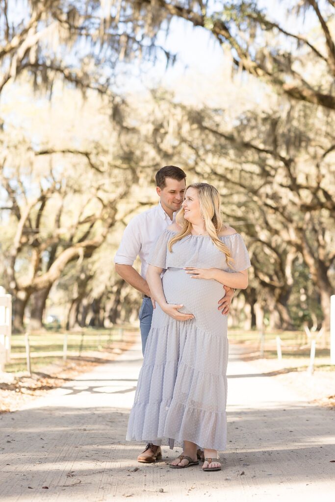 Gorgeous spanish moss trees make the perfect backdrop for a Savannah maternity session at Wormsloe Historic Site with Wisp + Willow Photography Co.  Mom wears a grey, eyelet dress and dad wears a white button down shirt and grey pants to coordinate!