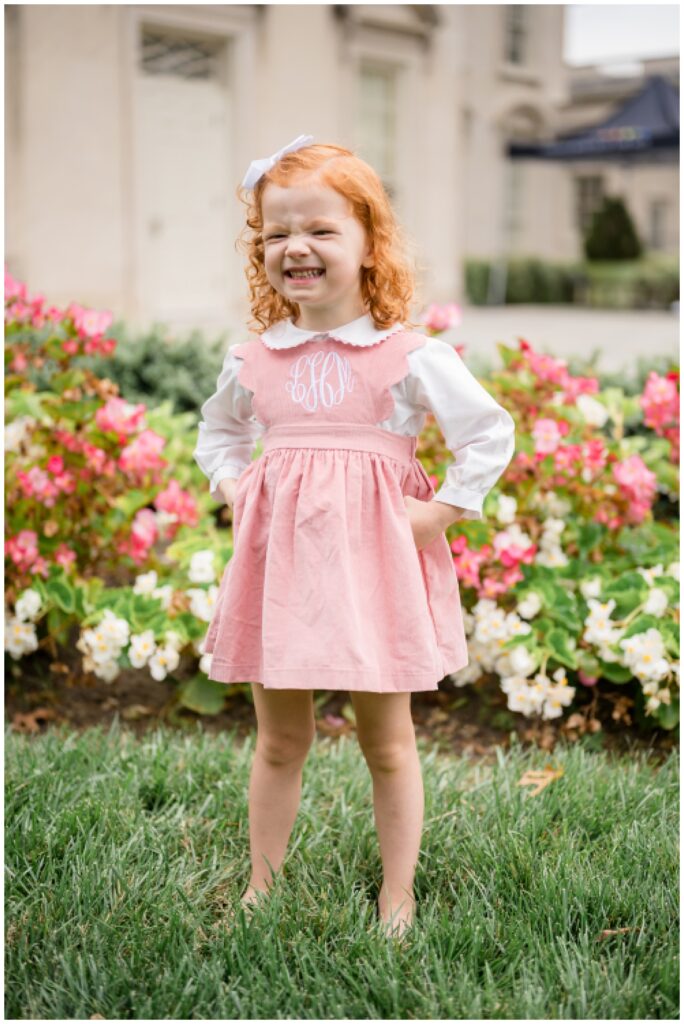 Little red head girl wears a white bow in her hair and a pink monogrammed dressed as she stands in the grass at the VMFA in front of the pink and white azalea bushes.