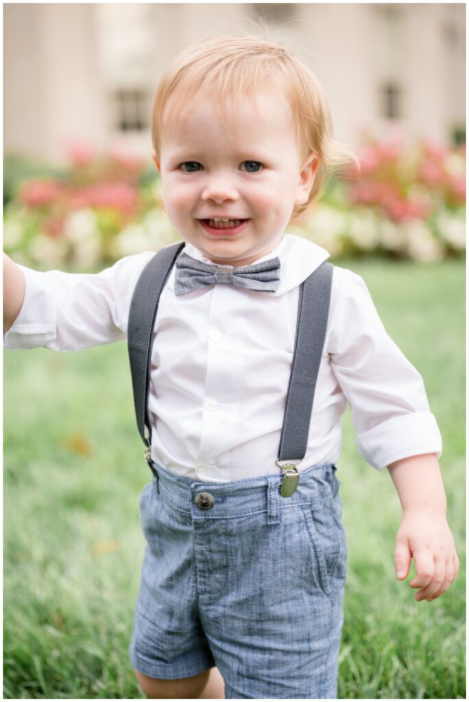 Little boy wears white shirt with a grey boy tie, suspenders and denim shorts as he smiles at the camera running across the grass at the VMFA.