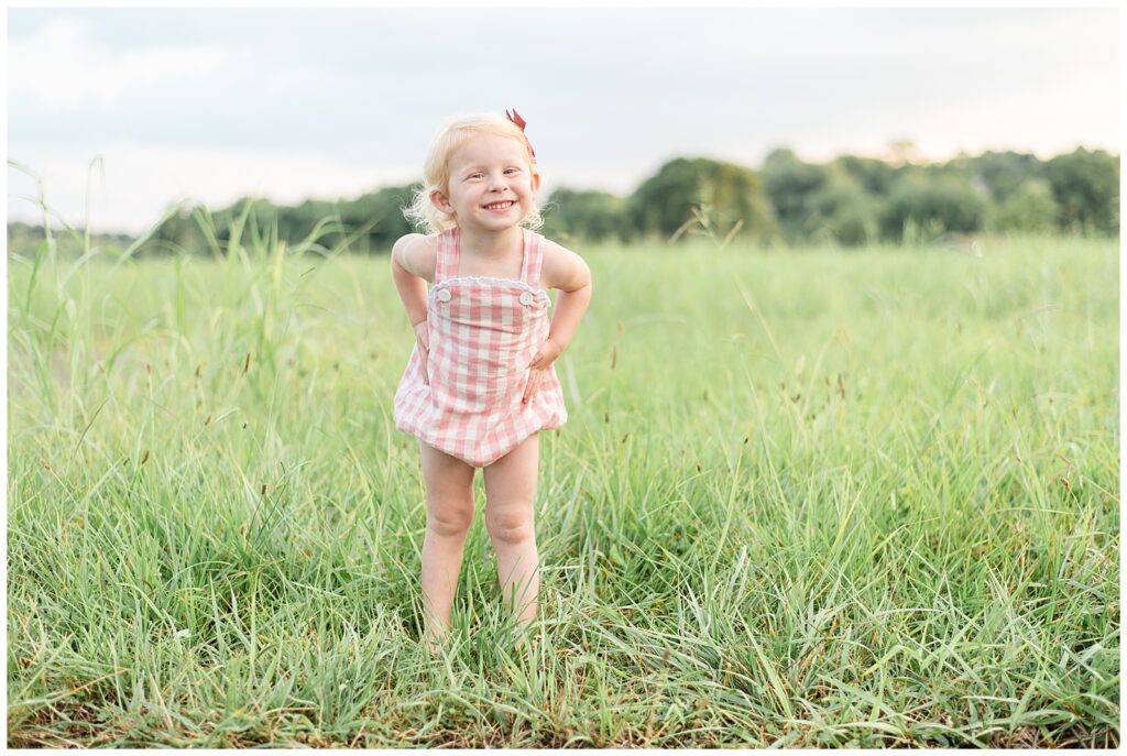 Little blonde hair girl stands in field at Cornelia Fort Airpark smiling at the camera and wearing a pink and white romper on a warm, sunny day.