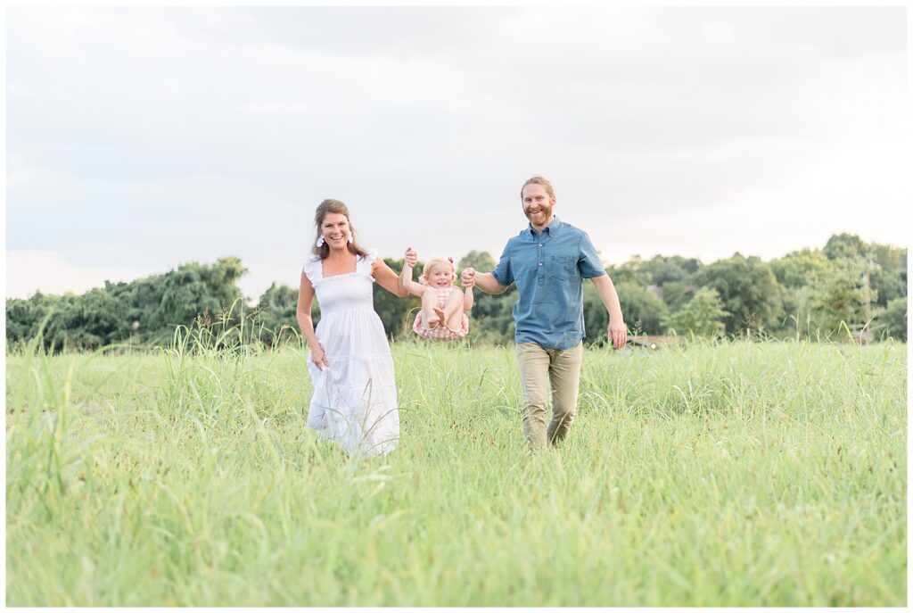 Mom, wearing a white long dress and Dad, wearing khakis and a blue shirt, swing their little girl who is wearing a pink and white romper through the fields of Cornelia Fort Airpark in Nashville, TN!