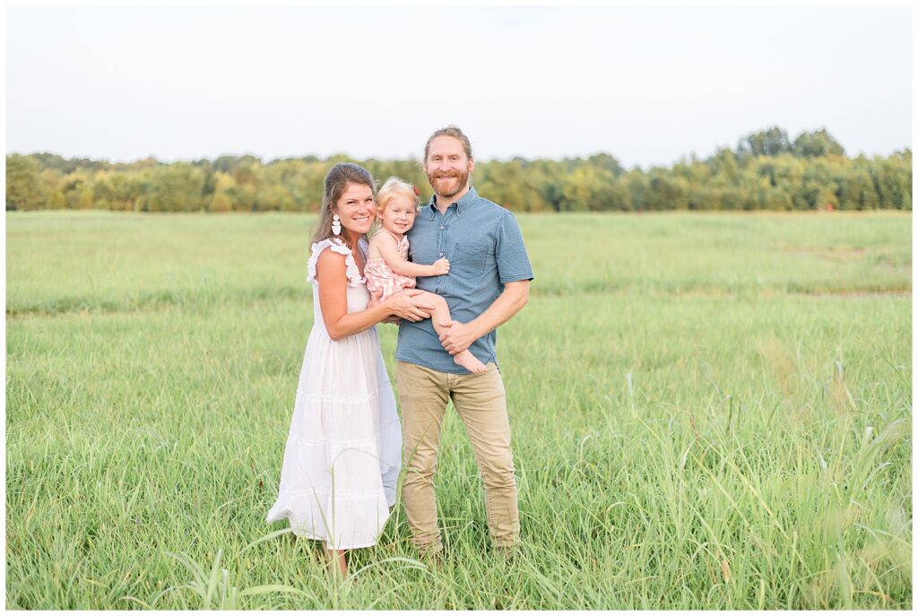 Family of three stand in a field of grass while Dad holds little girl on his hip and mom comes in close and holds her from behind as they smile at the camera of Wisp + Willow Photography Co. during their Nashville family session.