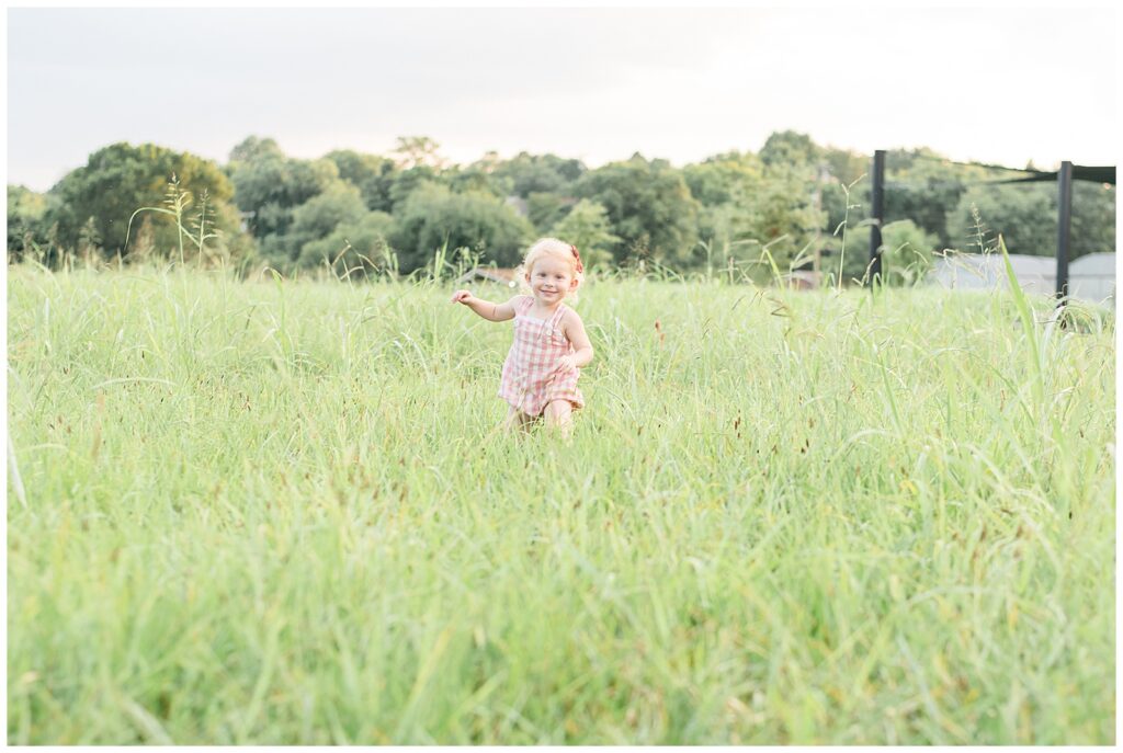Little girl wearing a pink and white plaid romper runs through the field of grass at the camera and smiles big as she is living her best life!