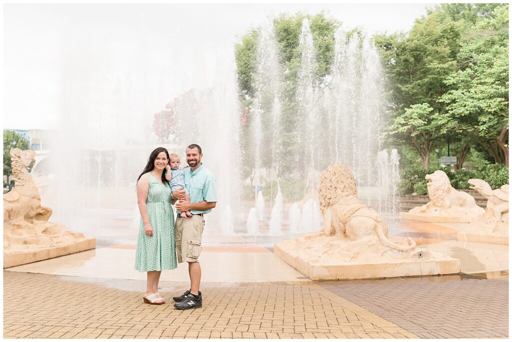 Family of 3 stand in front of the fountain at Coolidge Park in TN for their family session with Wisp + Willow Photography Co.