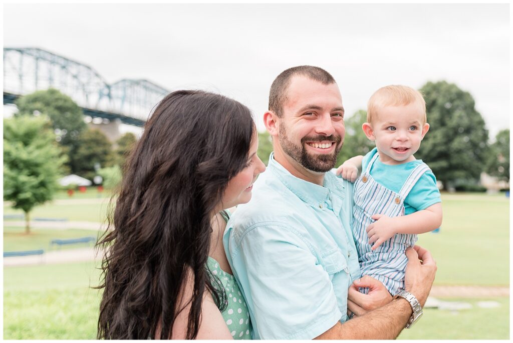 Dad holds toddler son and smiles at the camera as mom looks over her husbands shoulder at her son.  They coordinate wearing blue and green colors in their outfits.