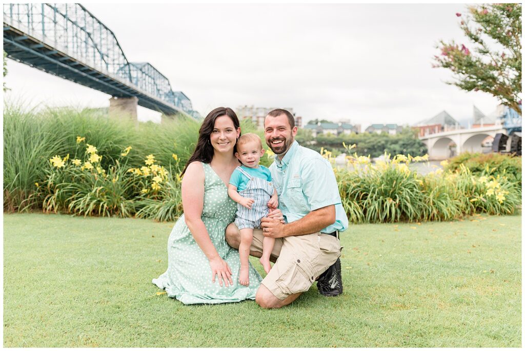 Chattanooga family photographer, Wisp + Willow Photography Co. capture family of 3 at Coolidge Park in TN in front of the yellow daylillies and bridge behind them as they kneel on the grass and Dad holds son on his knee.