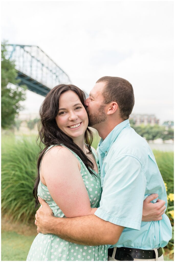 Husband and wife share a moment during their family session at Coolidge Park as they pull each other in close and husband kisses his wife on the cheek as she smiles at the camera.