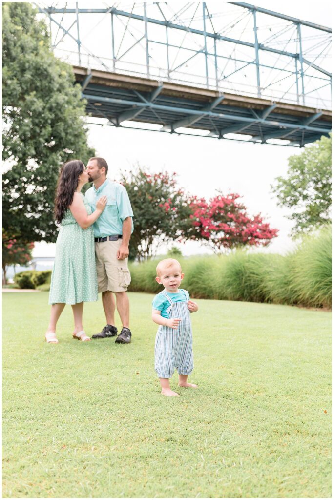 Wisp and Willow Photography Co, Chattanooga family photographer, captures toddler son in the forefront standing in the grass as his parents sneak a kiss behind him.