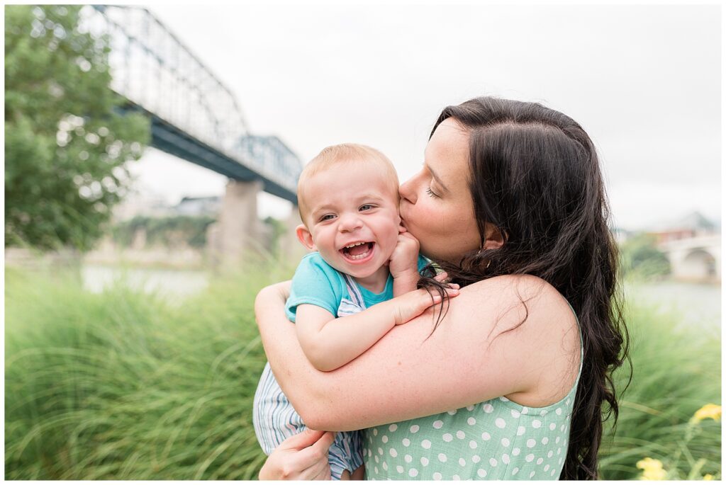 Mom kisses toddler son's cheek as he laughs just past the camera at his dad.  They stand in the grass at Coolidge Park in TN with the bridge and water behind them.