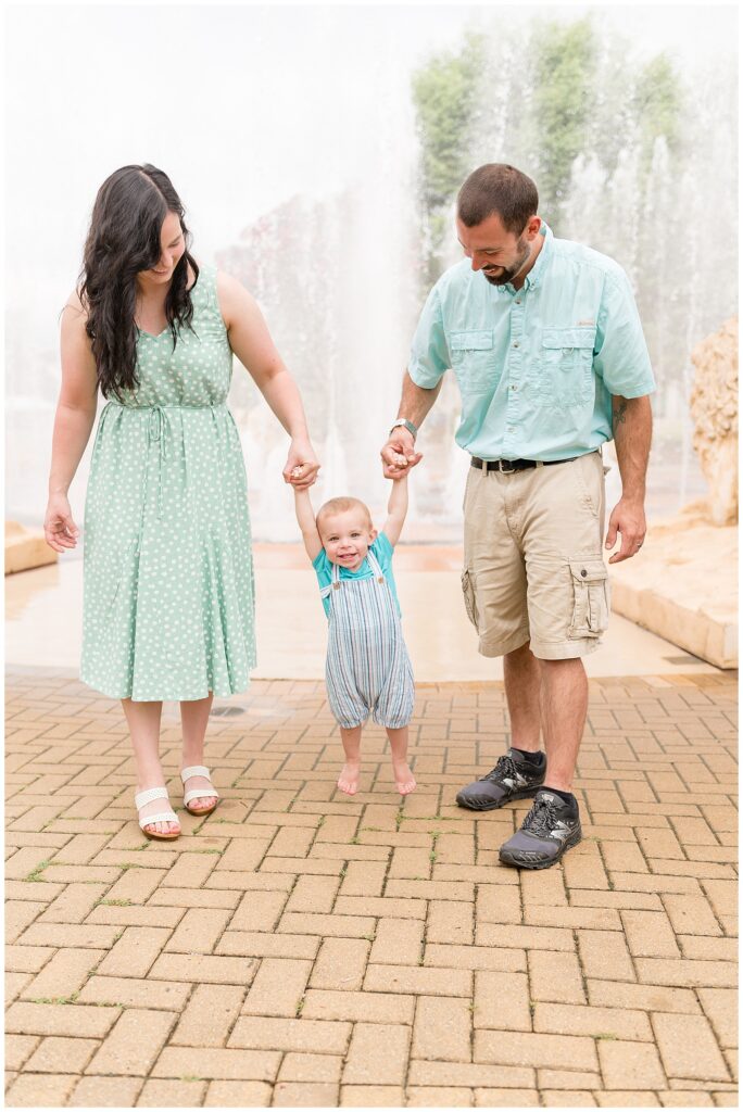 Mom and Dad holds hands of toddler son as they swing him in front of the fountain at Coolidge Park in TN.  Check out the Wisp + Willow Photography Co. blog to see more of this sweet family!