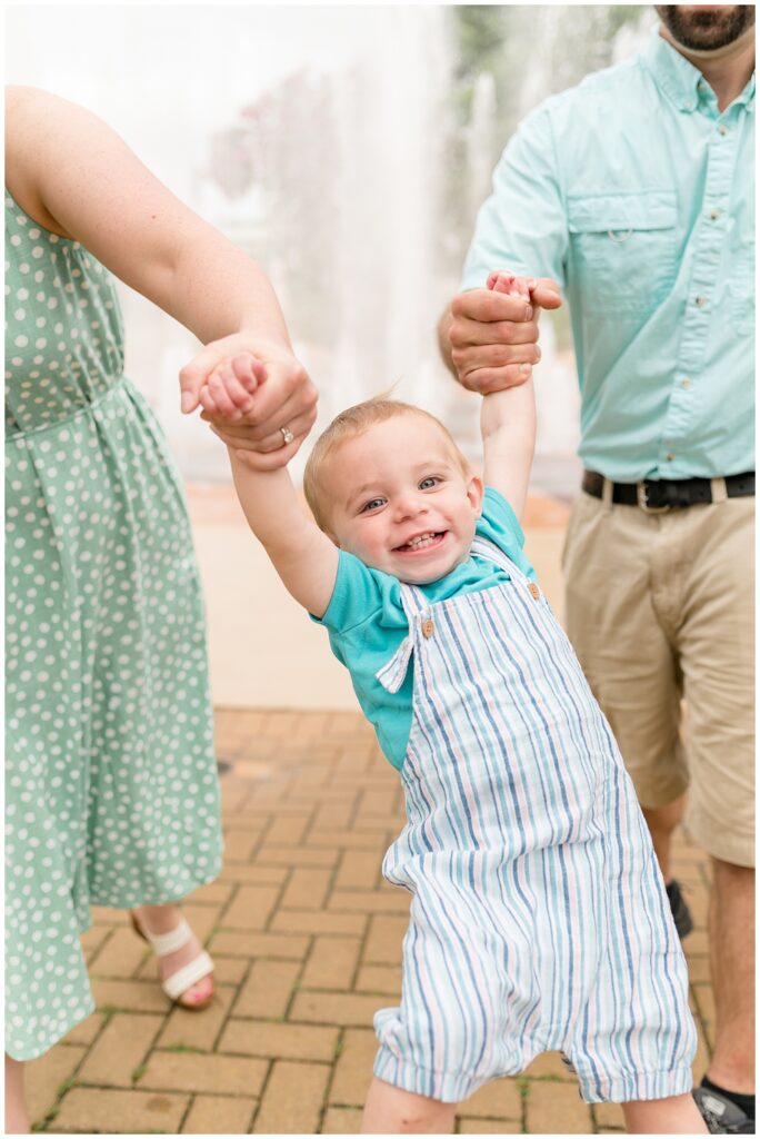 Toddler boy, who is wearing an aqua blue t-shirt and striped overalls, holds hands of parents as he is being swung and smiles the biggest smile at Chattanooga family photographer, Wisp + Willow Photography Co.