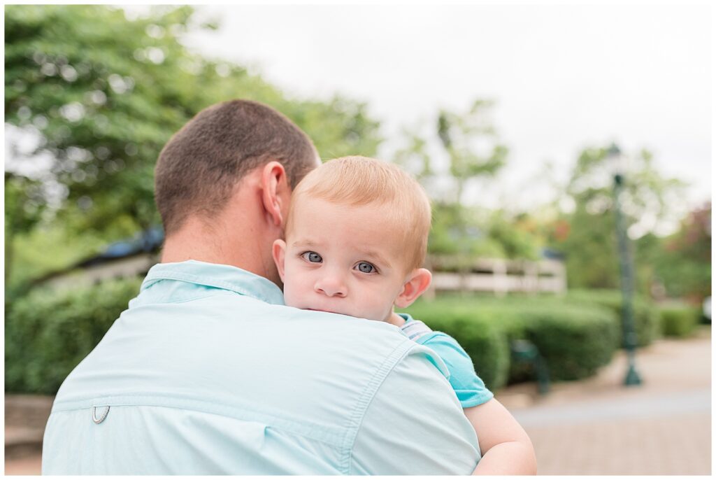 Toddler son holds on to Dad and looks over his shoulder at the camera of Wisp + Willow Photography Co. with sweet, sleepy, blue eyes.