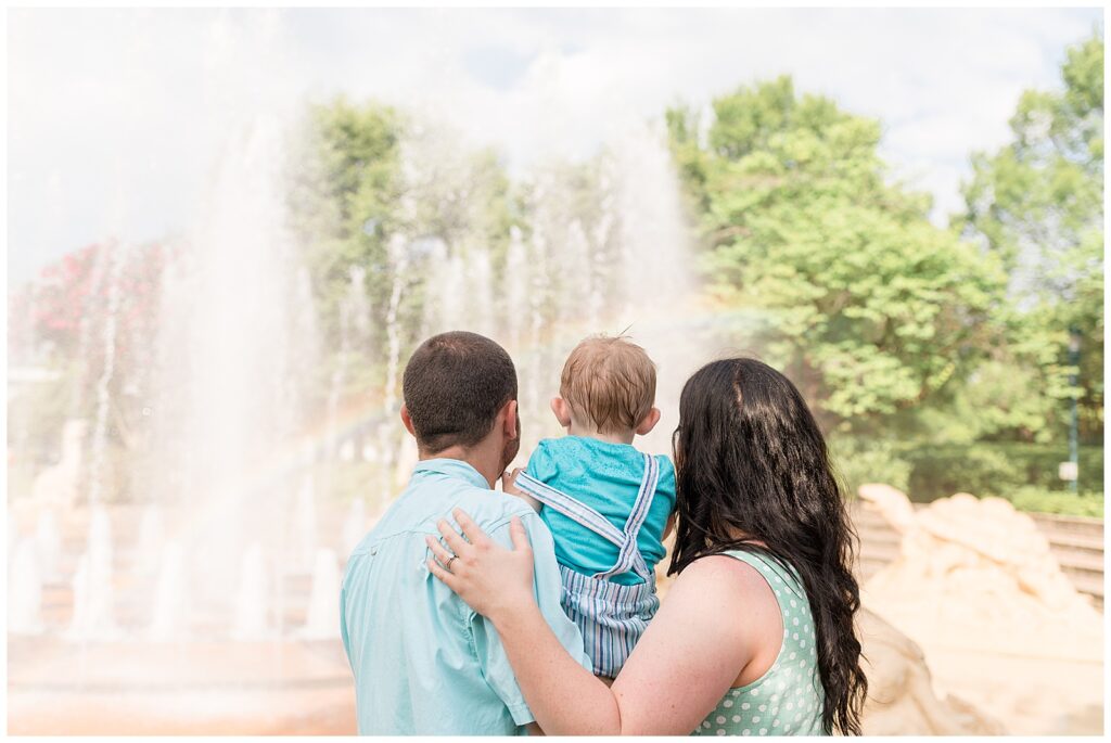 Mom, Dad, and toddler son, coordinating in blue and green outfits, face away from the camera as they look at the fountain in Coolidge Park and see a rainbow being made from the water and sun!