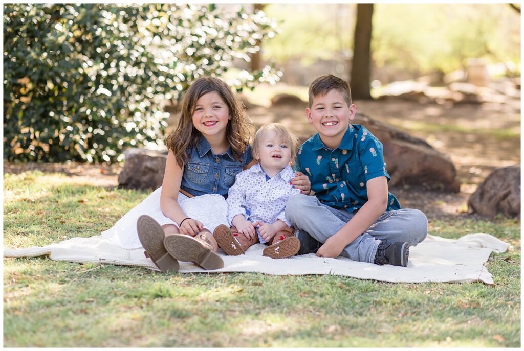Oldest brother wears a pineapple blue shirt and jeans, sister wears a denim sleeveless shirt and a white lace skirt, while youngest brother wears brown pants and a white shirt for their their Frisco family session at Central Park in TX.