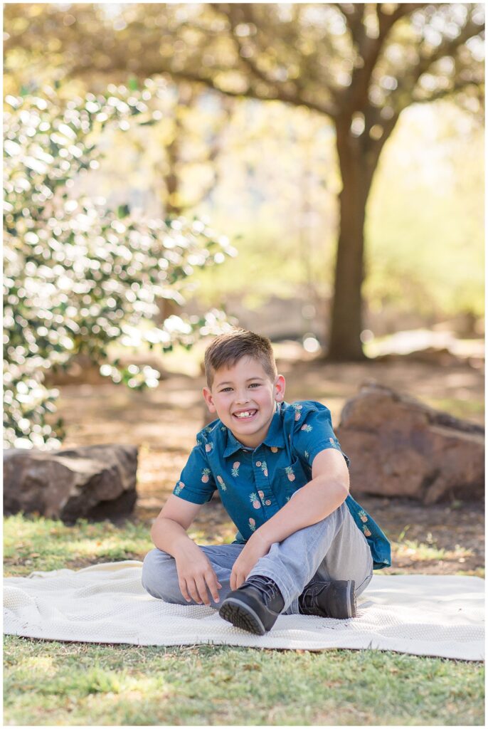 Individual picture of young boy as he sits on a textured cream blank as he sits with one leg tucked and the other bent as he smiles at the camera of Wisp + Willow Photography Co.