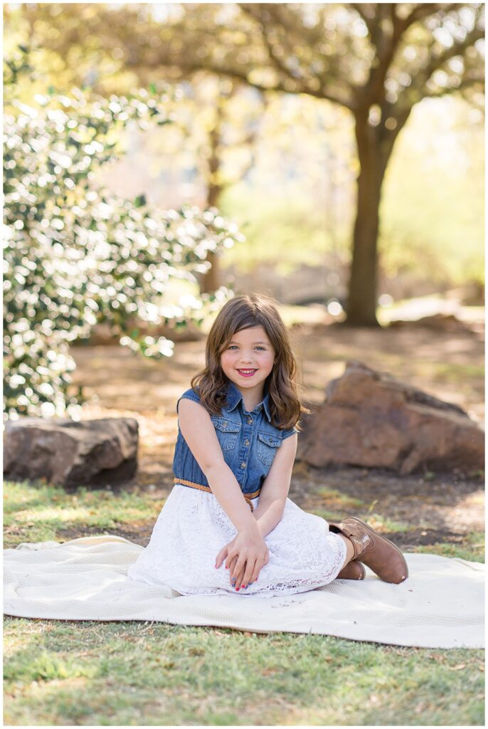 Young girl sits on textured cream blanket at Central Park in Frisco TX, wearing a lace white skirt, cowgirl boots, and a denim sleeveless shirt as she smiles at the camera.