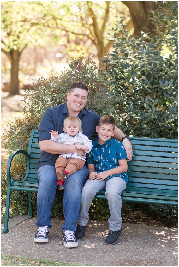 Dad sits on a green bench with his two young sons as they smile at the camera of Wisp + Willow Photography Co. during their Frisco family sesison.