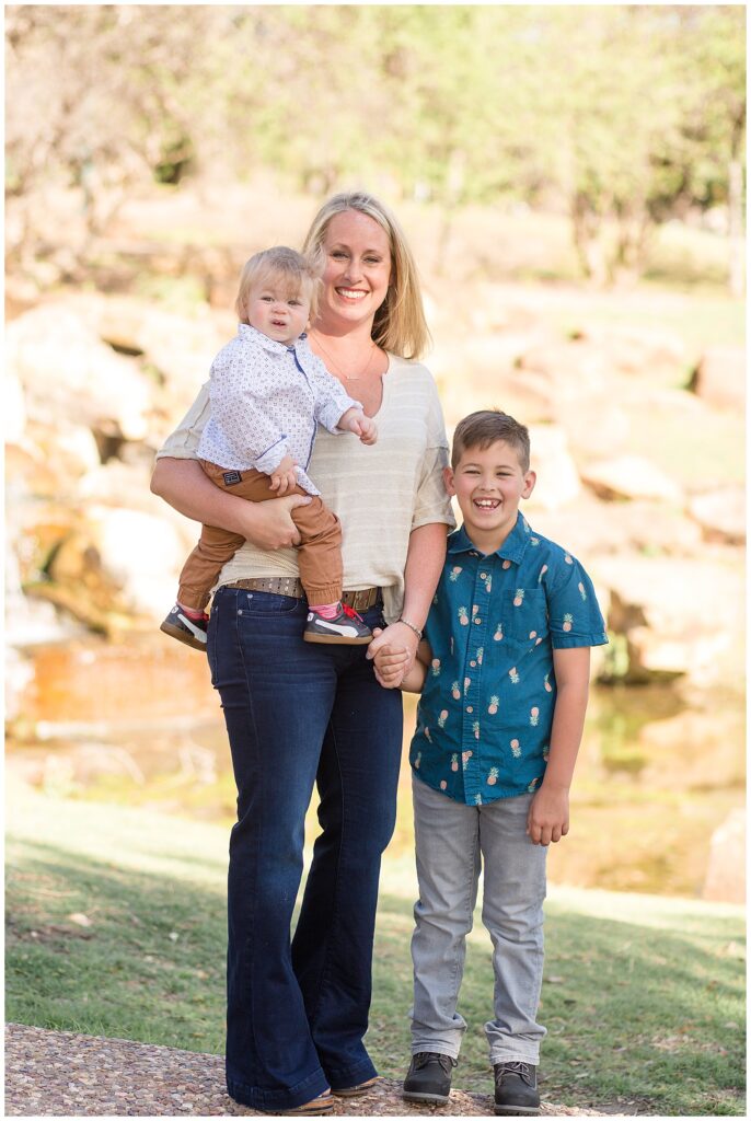 Mom stands holding hands with young son and holds her toddler son on her other hip as they smile at the camera of Wisp + Willow Photography Co.