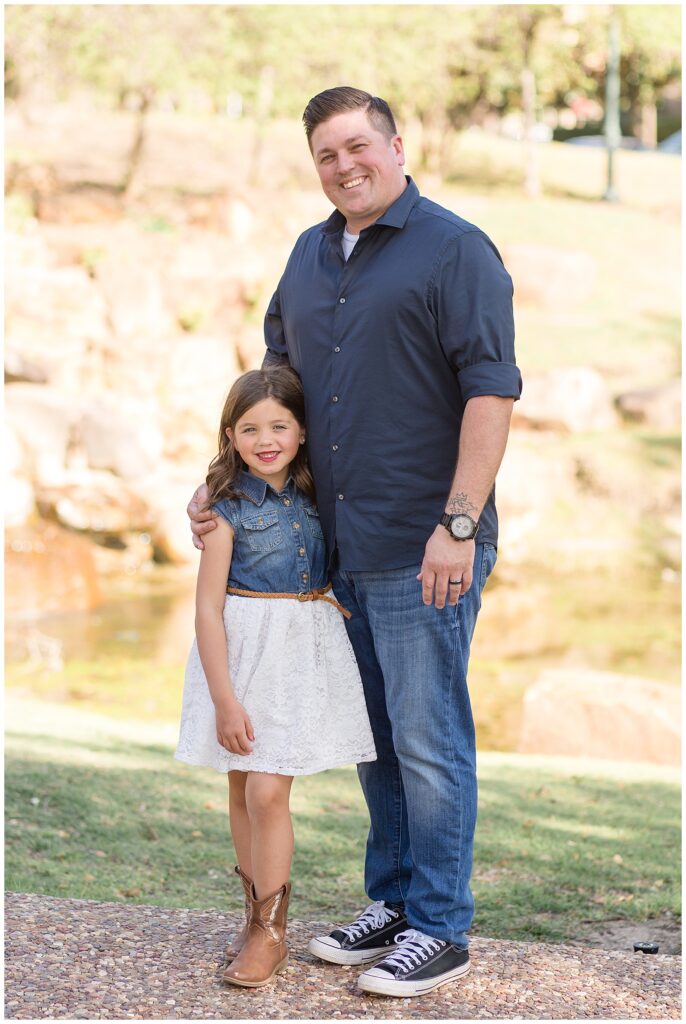 Dad stands as his daughter hugs his leg and they smile at the camera of Wisp + Willow Photography Co. during their Frisco family session.