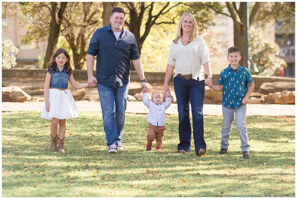 Family of 5 stand in a line holding hands and walk towards the camera of Wisp + Willow Photography Co. during their Frisco family session at Frisco Central Park.  