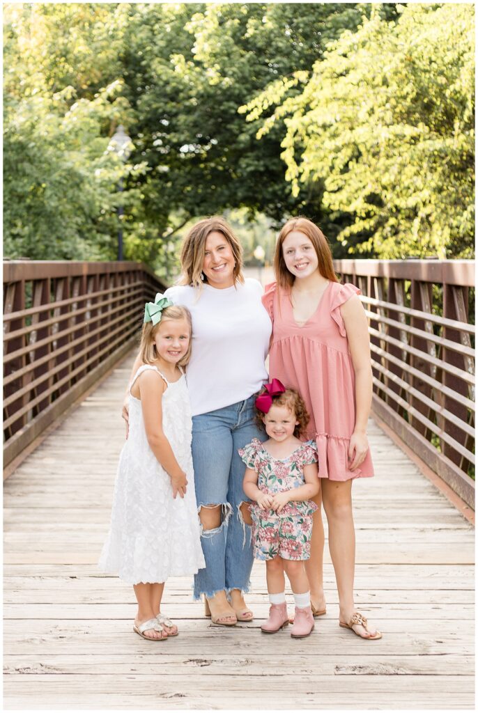 Mom, who wears a white tshirt and ripped jeans hugs oldest two daughters, one who wears a blush dress and the other who wears a white dress and the youngest stands in front wearing a floral outfits with a mix of blush, white, blue and green colors.