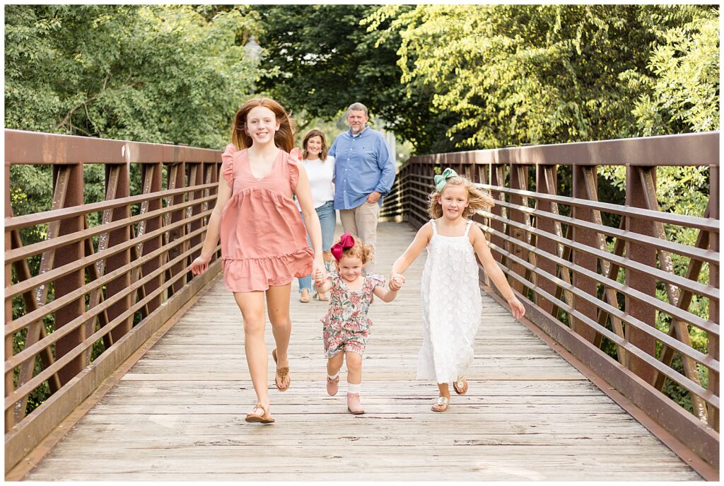 Mom and Dad stand in the background as their 3 daughters hold hands and run across the bridge towards the camera of Wisp + Willow Photography Co.