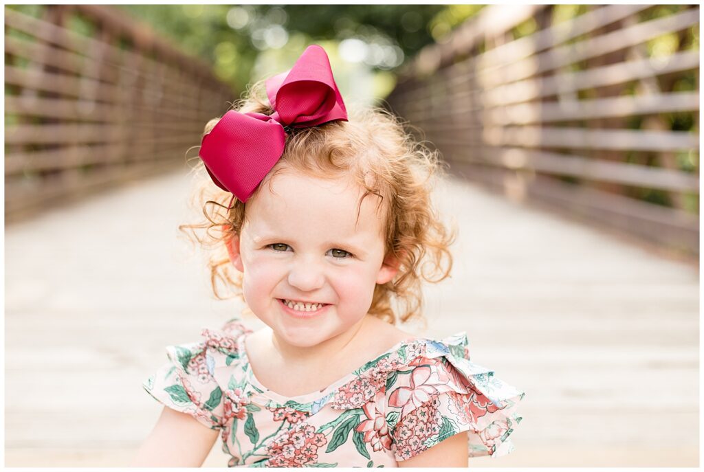 Little girl sits on a bridge at Pinkerton Park in Franklin, Tennessee and smiles at the camera with a burgundy bow in her red, curly hair and floral top.