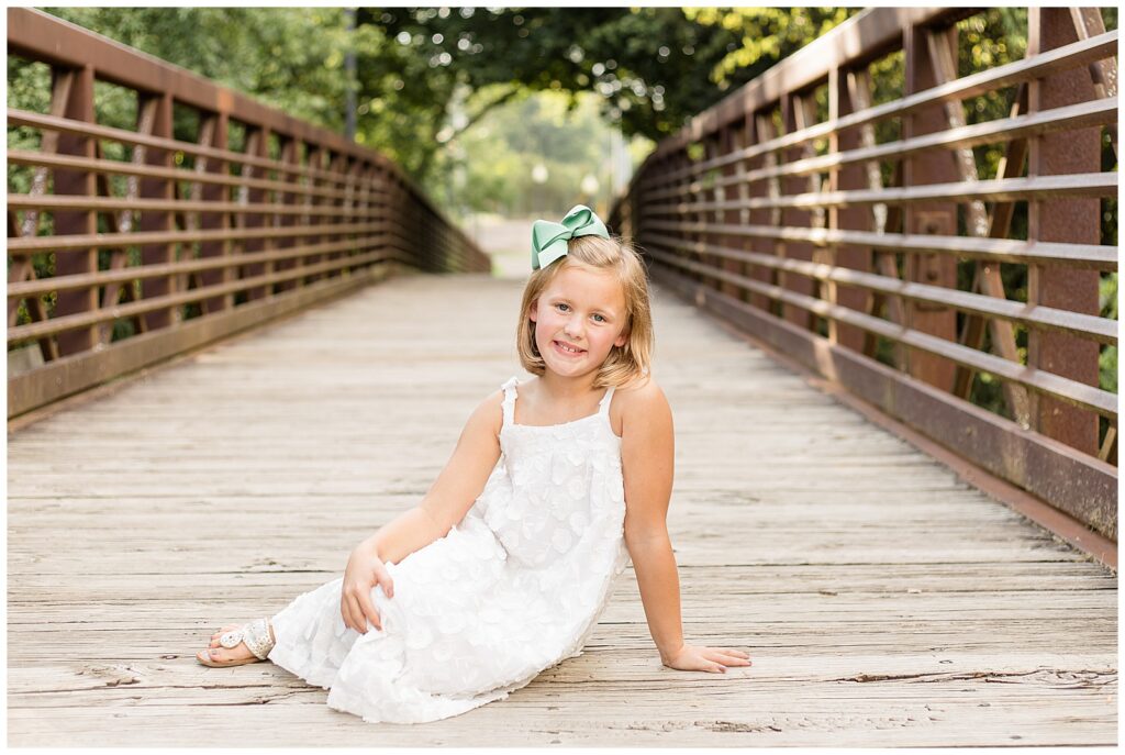 Young girl sits on a bridge with her legs swung to the side and smiles at the camera of Wisp + Willow Photography Co. with her blonde hair with a mint bow in it and a white, spaghetti strap dress.