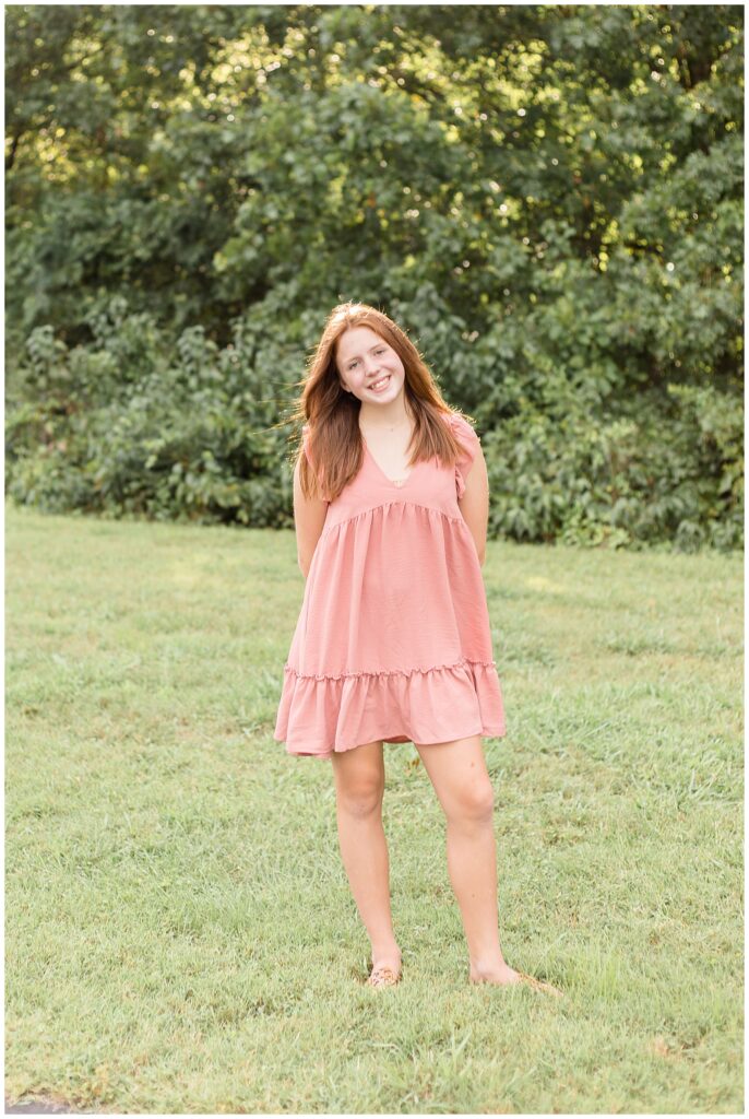 Young teenage girl, with red hair, stands in the grass with trees behind her, wearing a blush dress smiles at the camera of Wisp + Willow Photography Co. during their Franklin family portraits.
