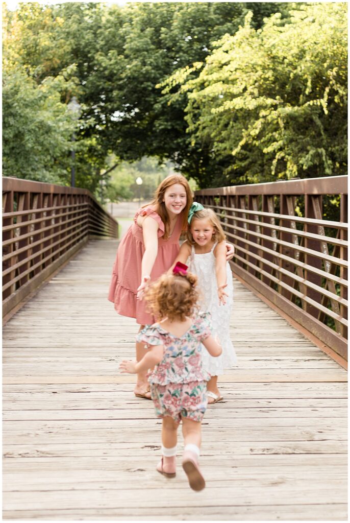 Two older sisters stand on the bridge at Pinkerton Park in Franklin, TN holding out their arms with the biggest smiles as their little sister with red, curly hair funs back towards them.
