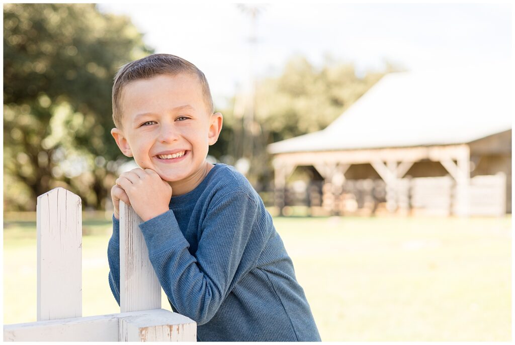 Young boy stands on a white fence and rests his hands on a post and his check on his hands as he smiles at the camera during his Texas family portrait session.
