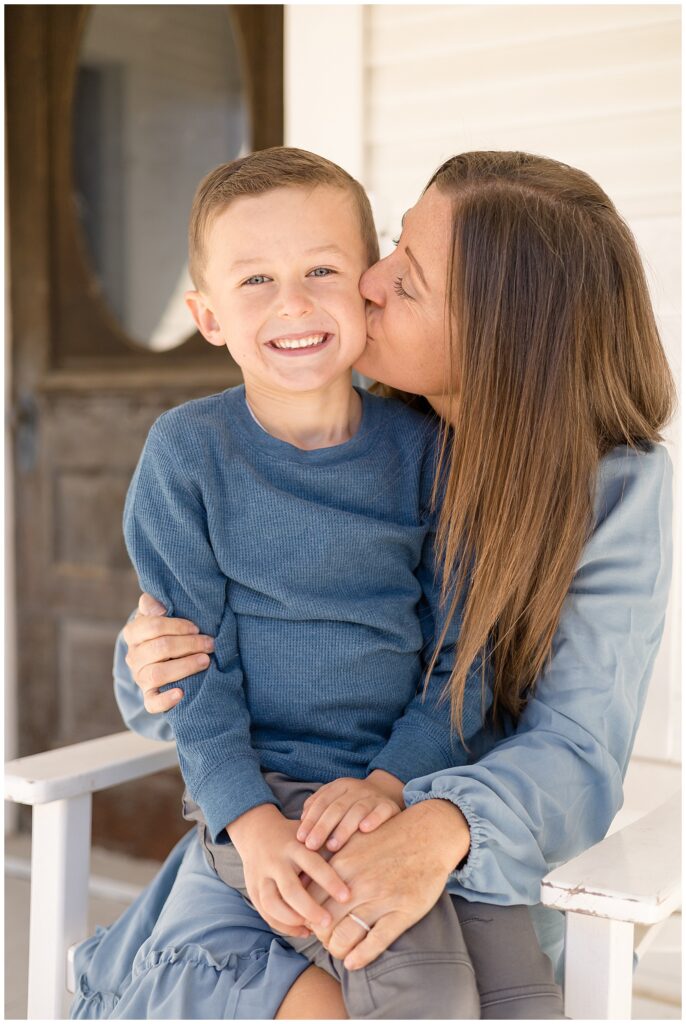 Mom sits in white rocking chair as her young son sits on her lap and she gives him a big kiss on his cheek as he smiles for his picture with mom.