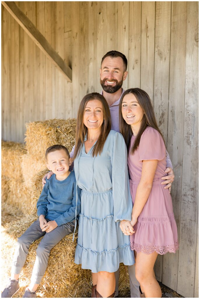 Family of 4 stand near the hay barrels with mom and daughter wearing dresses and dad and son wear long sleeves and slacks.