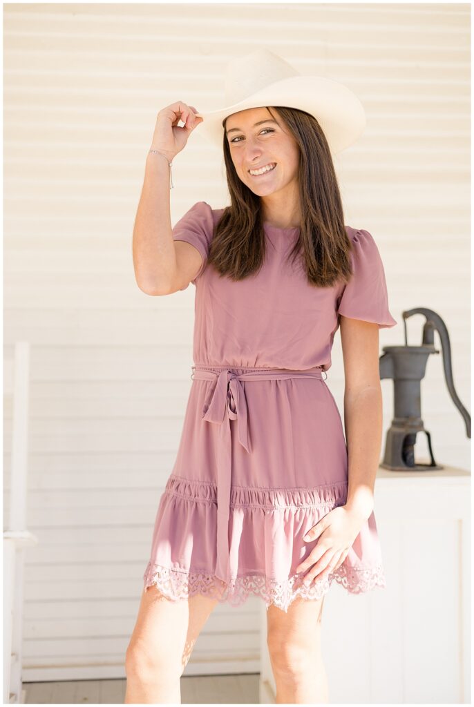 Teenage girl wears a blush colored dress and puts her hand up to her white cowgirl hat and smiles at the camera with a country feel.