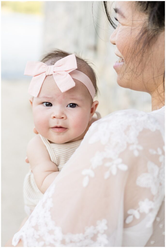 Wisp + Willow Photography Co. capture baby girl who is wearing a white and khaki thin stripe romper and a pink bow headband as she smiles over her mom's shoulder.  Click to see more on the blog today!