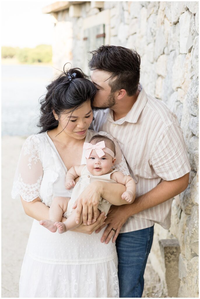 Family of 3 stand in front of a cobble stone wall as the husband gives his wife a kiss on her temple and she looks down smiling at her baby girl that she is holding.