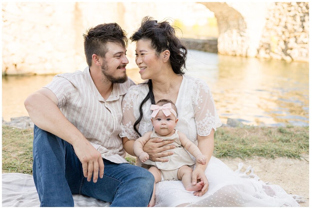 Family of 3 have an Adriatica family session in McKinney, TX.  They sit in front of the water as husband and wife look at each other and their baby girl looks at the camera of Wisp + Willow Photography Co. as she sits on her moms lap.