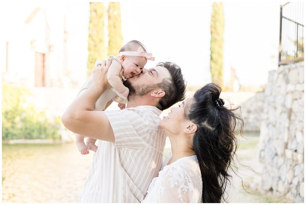 Family of 3 take family portraits at Adriatica Village as they coordinate with white lace and stripe outfits.  Baby girl smiles big and down at Mom as Dad lifts her up and gives her a kiss on her cheek.  Click to see more on the blog today!