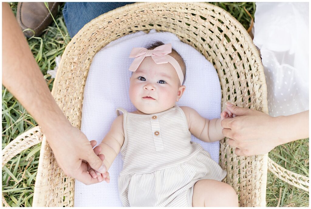 Baby lays in wicker, travel basket and looks up at camera with her pink, bow headband and holds the fingers of her parents.  