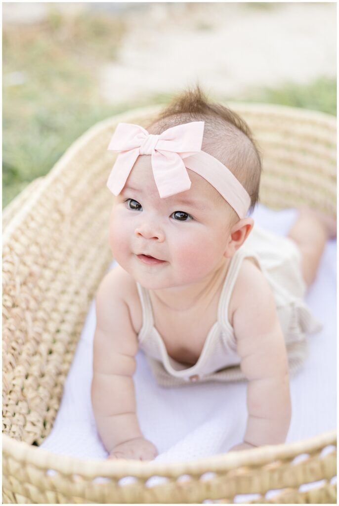 Adriatica family session in McKinney, TX has a baby girl laying on her belly and pushing up with her hands in a travel bassinet as she smiles off with big brown eyes and wears a pink bow headband.