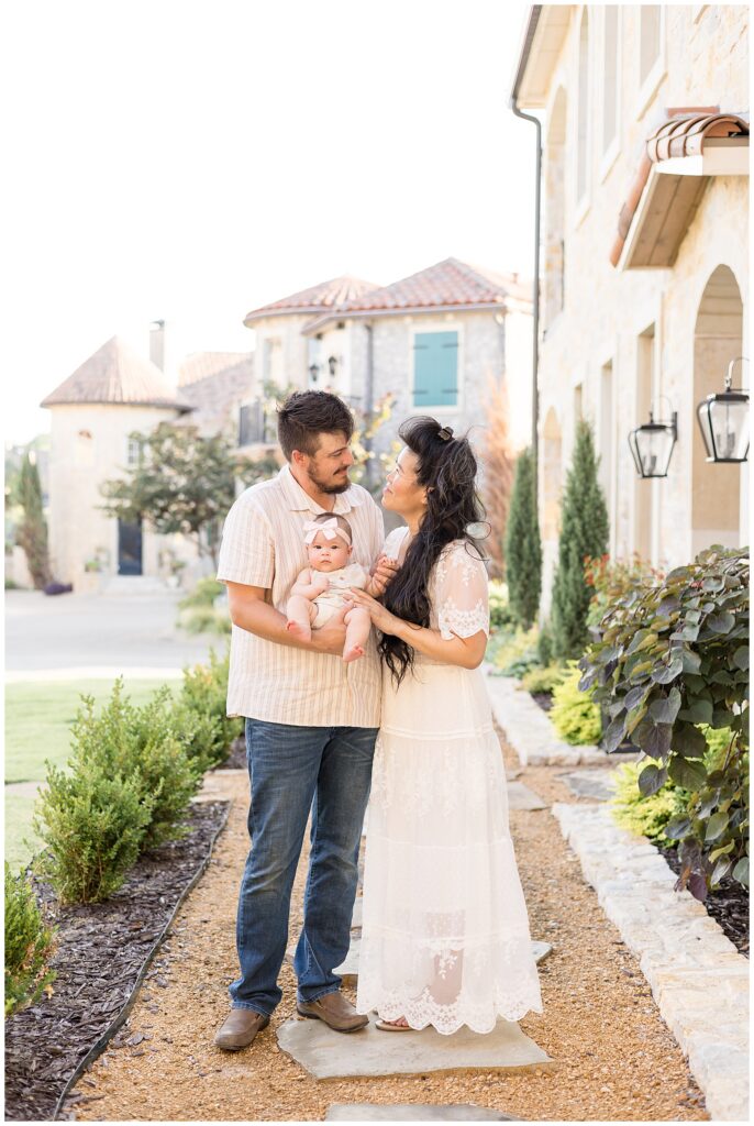 A family of 3 stand on the pathway at Adriatica Village as they hold their baby girl and look up at each other.  They coordinate with white and cream colors and a mixture of lace and stripe textures.
