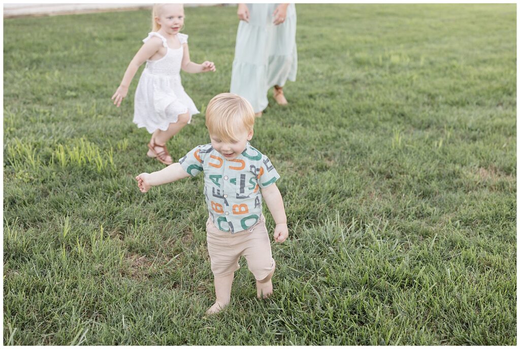 Blonde hair boy runs in grass wearing a button up letter shirt and khaki shorts as his sister in a white dress runs behind him.