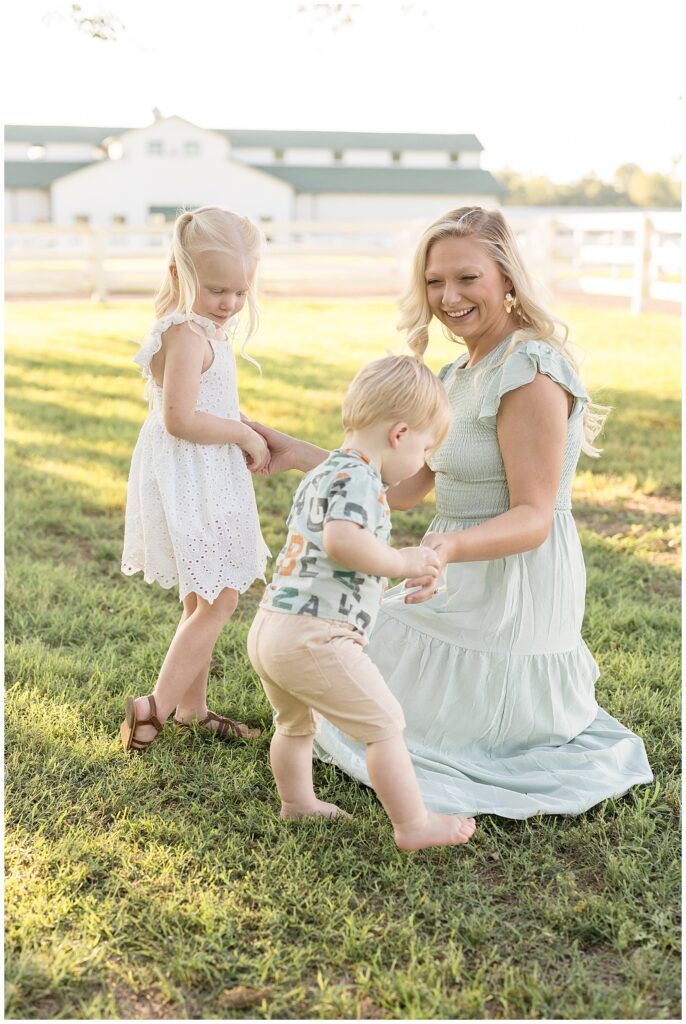 Mom who wears a mint green, long dress sits in the grass at Harlinsdale Farm as she holds the hands of her daughter and looks down at her toddler son.