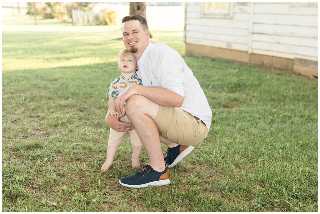 Father and son take a picture during their family session with Wisp + Willow Photography Co. as Dad squats down to the ground and his toddler son snuggles in close.