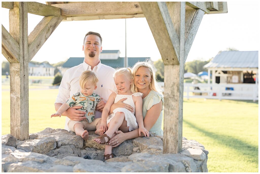 A family portrait at Harlinsdale Farm is captured by Franklin photographer, WIsp + Willow Photography Co.  The family of 4 with two young children sit on a well as Mom and Dad stand behind them and smile at the camera.