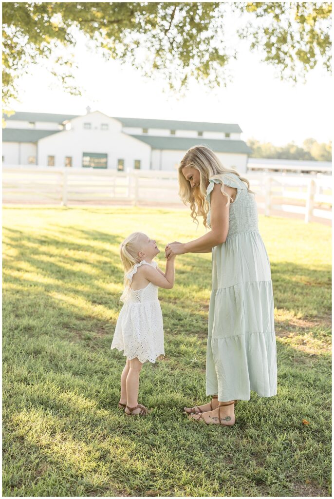 Mom who has blond hair and wears a mint green long, maxi dress holds her daughters hands as she looks down at her and they smile at each other.